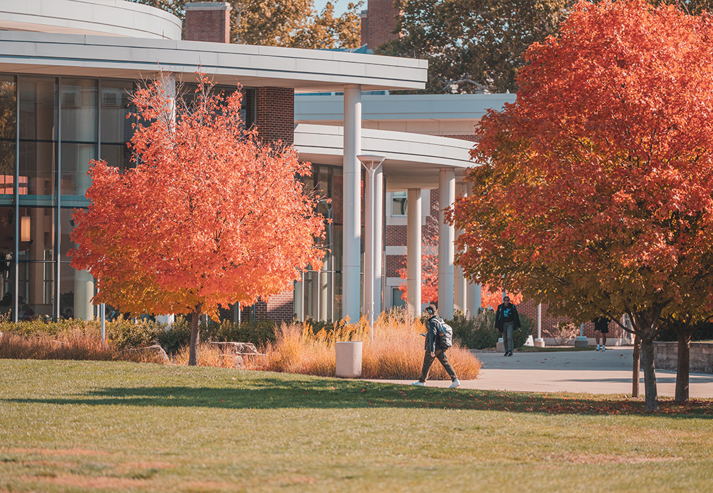 The photo shows an outdoor image with two students walking near the SDRP building in the fall, as the leaves on the trees are bright and colorful.