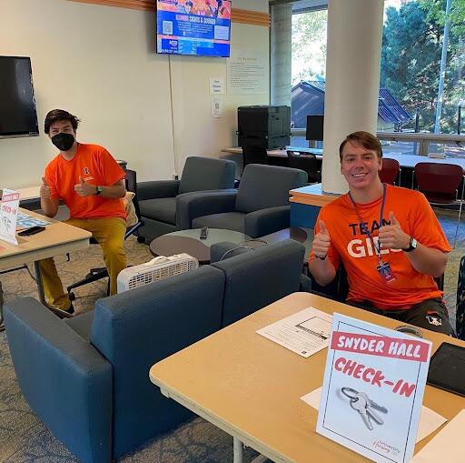 Two RAs in orange shirts sit behind check in tables with a thumbs up