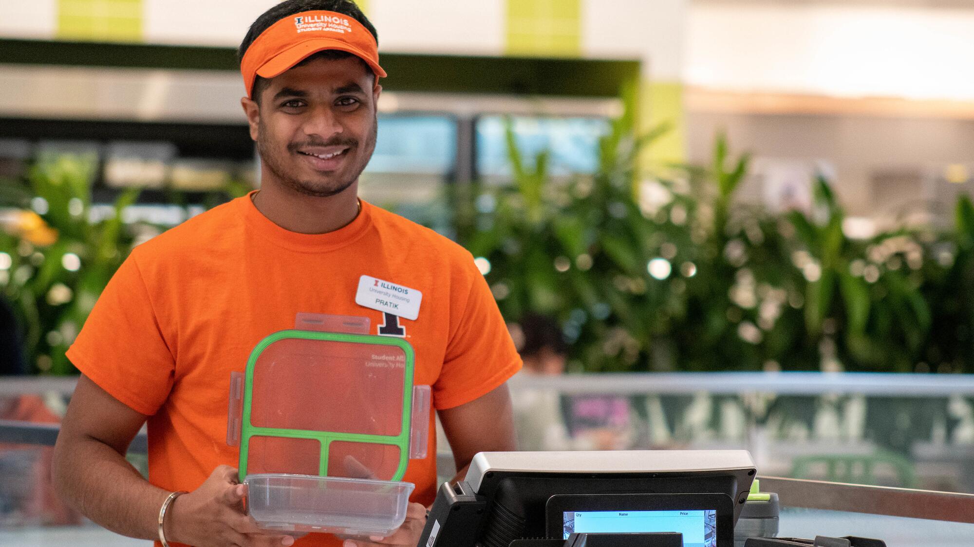 A University Housing employee smiles as he holds and displays a Good2Go container at a dining hall.